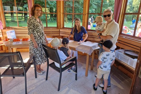 In einem Pavillon stehen zwei Frauen und zwei Kinder. Eine der Frauen hält ein Kleinkind im Arm. Auf einem Sessel sitzt ein Kind, auf dessen Schoß sitzt ein kleineres Kind. Durch die Fenster des Pavillons sieht man ins Grüne.