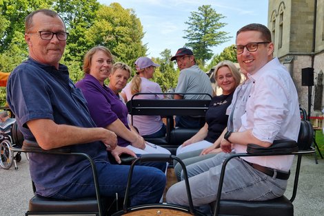Frauen und Männer sitzen in einer Kutsche, die sich in einem Park befindet. Der Kutscher wendet seinen Kopf zurück.