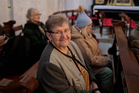 eine grauhaarige Frau sitzt mit anderen Menschen in einem Kirchengestühl