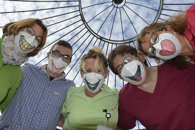 Fünf Männer und und Frauen stehen gebückt im Halbkreis nebeneinander. Sie haben Masken mit lachendem Mund über dem Mund. Ihre Augen sind zusammengekneift.