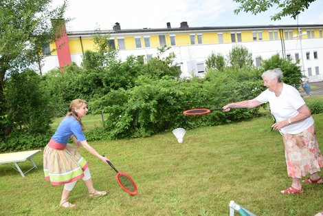 Zwei Frauen spielen in einem Garten Federball. Es ist ein Teil einer Liege zu sehen und im Hintergrund ein langgestrecktes weiß-gelbes Wohnhaus.