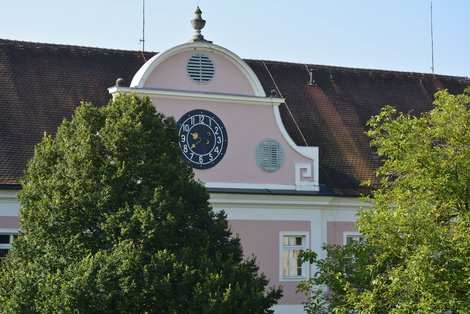 Eine barocke Dachbodenfront mit großer schwarzer Turmuhr. Davor zwei Bamkronen.