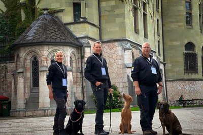 Eine Frau und zwei Männer, alle dunkel gekleidet, stehen lächelnd mit ihren drei Hunden vor einem Schloss.