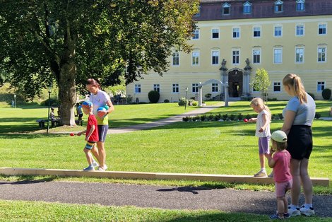 Zwei Kinder balancieren mit je einem Löffel, auf dem sich ein Ei befindet über einen Balken, der sich auf einer Wiese mit einem Baum vor einem Schloss befindet. Eine Frau und ein kleines Mädchen beobachten die Szene.