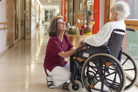 Eine weißhaarige ältere Dame sitzt in einem Rollstuhl. Vor ihr eine Pflegerin mit bordeaux-färbigem Kassak und weißer Hose in der Hocke. Hinter den beiden ein Tisch mit einem Osterstrauß.