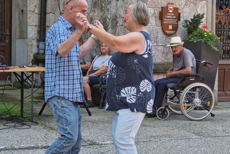 Eine Frau mit blauer kurzärmeliger Bluse mit großen weißen Blättern tanzt mit einem Mann, der ein blau-kariertes Hemd trägt vor einem Haus. Im Hintergrund stehen ein Mann im Rollstuhl und eine Frau zu. 