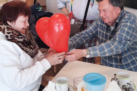 Ein Mann mit kariertem Hemd schenkt einer Frau im weißen Pullover einen roten herzförmigen Luftballon. Beide lächeln.